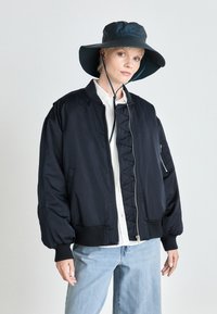 Marine Bomberjacke mit gerippten Bündchen und Saum, ausgestattet mit einem Reißverschluss vorne und seitlichen Taschen. Kombiniert mit einem breitrandigen grünen Bucket Hat.
