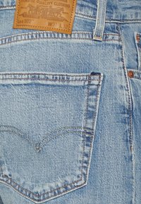 Vaqueros de denim azul claro con un bolsillo trasero que presenta un patrón de costura en forma de "V". La etiqueta de cuero en la cinturilla dice "Ropa de Calidad Genuina."