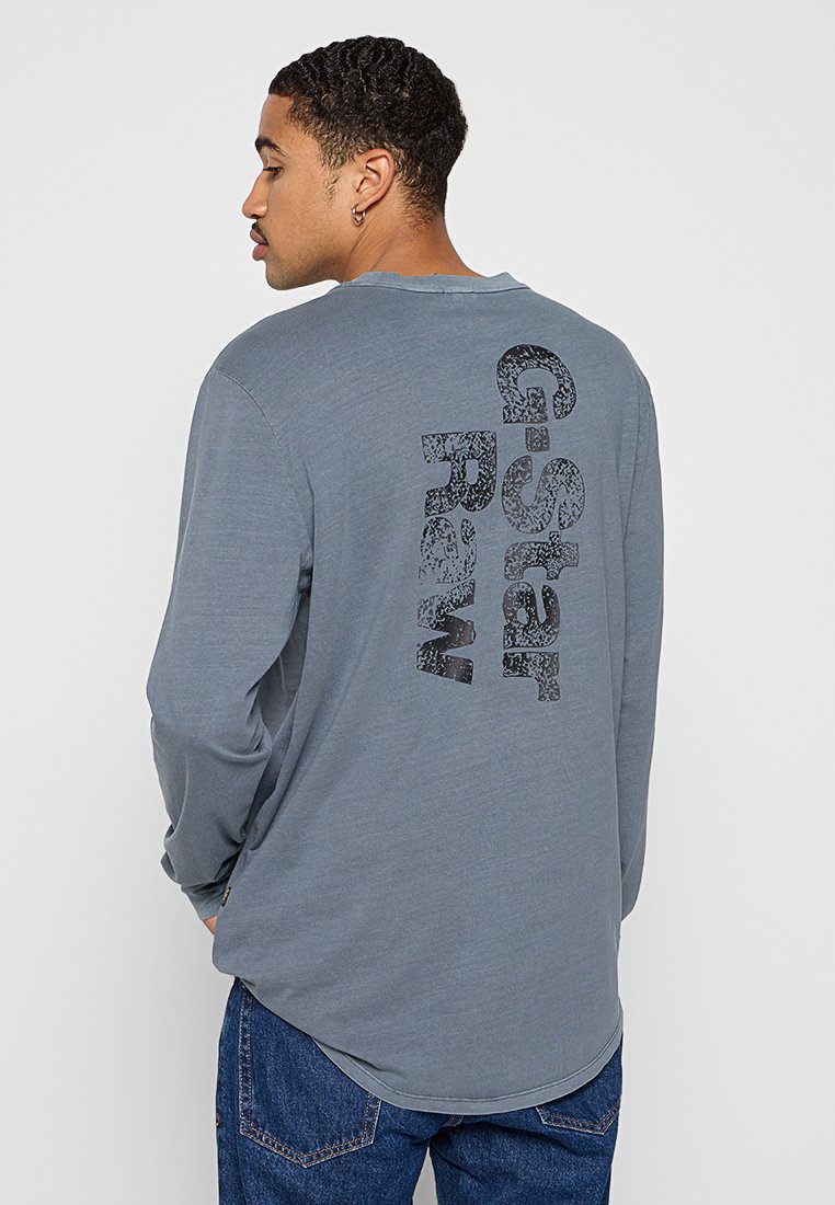 Mann von der Seite stehend, trägt ein graues Langarmshirt mit großem, vertikal auf dem Rücken aufgedrucktem "G-Star Raw"-Text, kombiniert mit blauen Jeans.