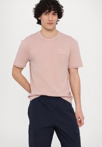 Lyserød bomulds t-shirt med rund hals og korte ærmer; har et tekstureret logo patch over venstre bryst. Modellen bærer navy cargobukser.