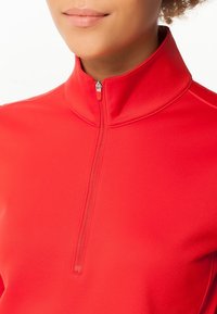 Primer plano de una persona que lleva una chaqueta roja con cremallera y un cuello alto, mostrando los labios y el cuello sobre un fondo blanco.