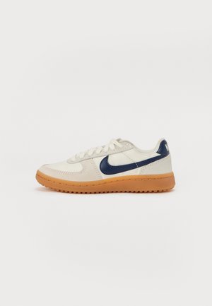 Sneaker bianca e beige con swoosh Nike blu navy e suola in gomma color caucciù, vista dal lato esterno su sfondo semplice.