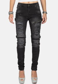 Jean skinny noir en denim, avec des accents déchirés, des détails usés et un petit patch logo sur la cuisse.