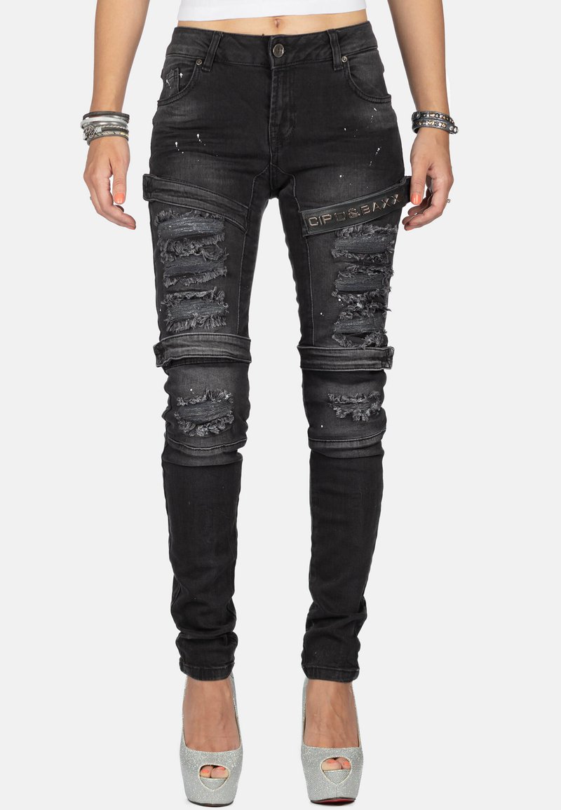Jean skinny noir en denim, avec des accents déchirés, des détails usés et un petit patch logo sur la cuisse.