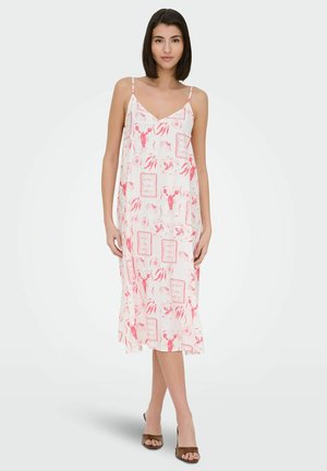 Femme debout portant une robe midi blanche sans manches avec des imprimés de homard rouge et fruits de mer et des talons ouverts marron sur un fond uni.