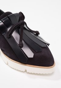 Mocassin en daim noir avec frange décorative en cuir noir et lacet noué, semelle intermédiaire beige et semelle extérieure blanche dentelée.