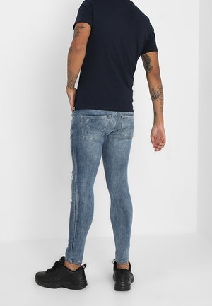 Homme portant un jean bleu moulant, un t-shirt noir et des baskets noires, debout dos au spectateur sur un fond blanc uni.