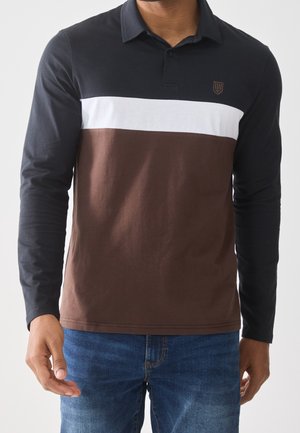 Mann trägt ein langärmliges Poloshirt mit schwarzen Ärmeln, weißem horizontalen Streifen, braunem unteren Teil und blauer Jeans, stehend vor schlichtem Hintergrund.