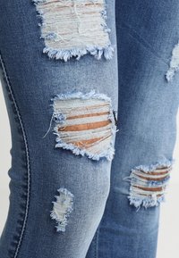 Verwaschene blaue Jeans aus Denim mit mehreren ausgefransten Rissen an den Knien, die einen helleren blauen Stoff darunter sichtbar machen; Slim-Fit-Design.