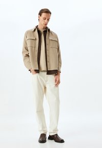 Beige button-up skjortejakke med to brystlommer, lagd over en brun skjorte, hvide jeans og mørkebrune læderstøvler.