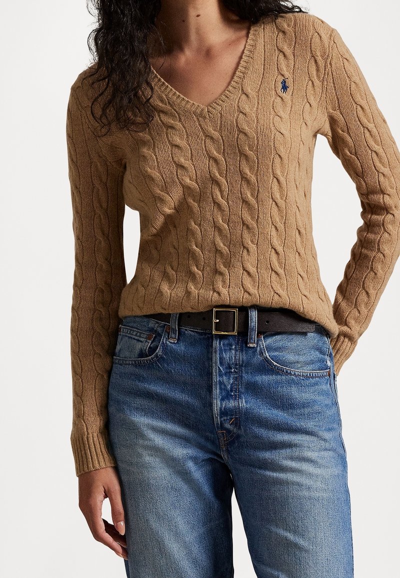 Femme portant un pull en maille torsadée couleur camel avec un col en V et un petit logo brodé ainsi qu'un jean bleu avec une ceinture noire.