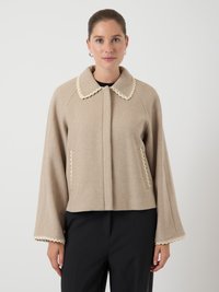 Béžová vlněná bunda se střihaným límcem a lemem, s roztřesenými rukávy a předními kapsami, nošena přes černé kalhoty na bílém pozadí.