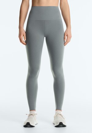 Osoba ubrana w szare legginsy z wysokim stanem i białe buty sportowe, stojąca na tle jednolitego jasnego tła.