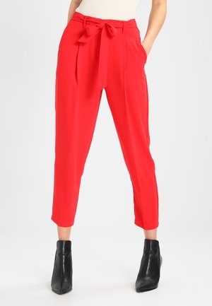 Pantalons rouges taille haute, plissés, avec une ceinture à nouer, dotés de poches latérales et d'une coupe raccourcie. Portés avec des bottines noires.