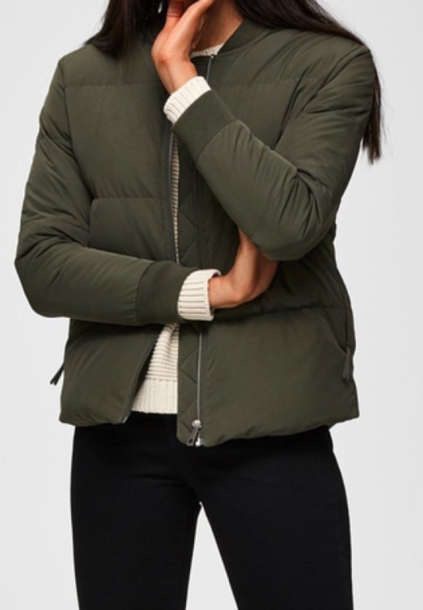 Giubbotto bomber trapuntato verde oliva con polsini a costine, tasche con zip e chiusura frontale con zip, indossato sopra a un maglione color crema.