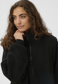 Sort fleecetakjakke med høj krave, lynlåslukning og oversized ærmer. Blød tekstur med en blød finish.