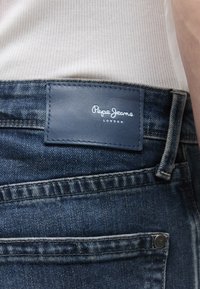 Primo piano della tasca di jeans di denim blu con etichetta in pelle blu scuro con la scritta "Pepe Jeans London" cucita sopra.