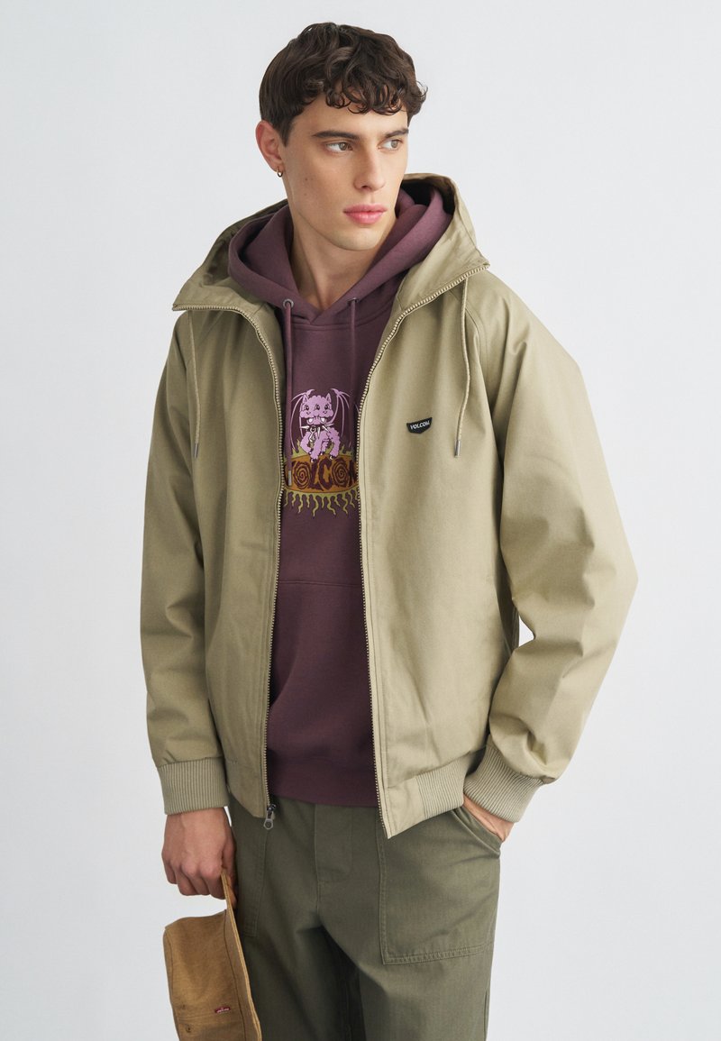 Χακί μπουφάν με φερμουάρ και κουκούλα, φορεμένο πάνω από μωβ φούτερ με τύπωμα, συνδυασμένο με πράσινο παντελόνι cargo.