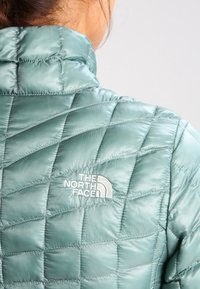 Světlá tyrkysová prošívaná bunda s vysokým límcem a bílým logem The North Face na levém rameni. Saténová textura a vzor v diamantovém tvaru.