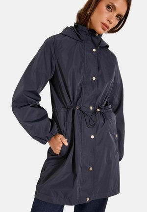 Kvinde iført en navy hættetrøje regnfrakke med trykknapper og snor i taljen, hænderne i lommerne, poserende mod en ensfarvet baggrund.