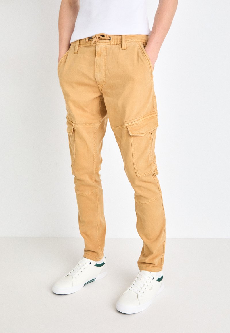 Beige cargobyxor med sidofickor, dragsko i midjan och en slim fit. Kombination med vita sneakers med gröna detaljer.