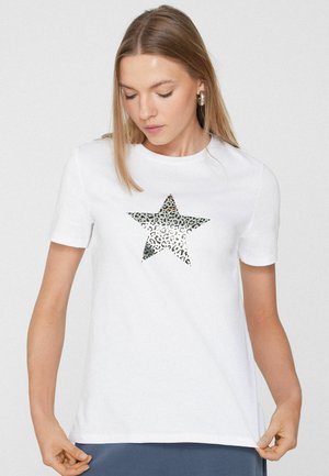 Kobieta w białej, krótkiej koszulce z błyszczącą gwiazdą w panterkę, patrząca w dół, trzymająca dół koszulki, stojąca na tle jednolitego, jasnego tła.