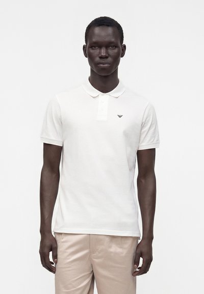 Modèle masculin portant un polo blanc à manches courtes avec un petit logo sur la poitrine et un pantalon beige, debout devant un fond blanc uni.