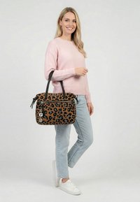 Naeratav naine, kandes roosat kampsunit ja heledaid teksaseid, hoides pruuni leopardimustriga kotti mustade sangadega ning seljas valged tossud.