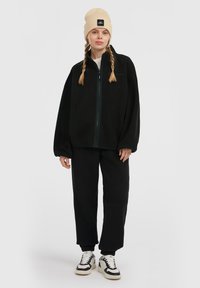 Black, fleece zip-up jacket with a high collar and pockets, paired with matching fleece pants. Beige knit beanie on top, and black-and-white sneakers.