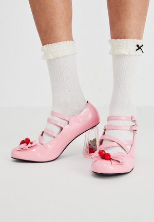 Tacchi in vernice rosa con punta arrotondata, tre cinturini e decorazioni a forma di ciliegia. Il tacco a blocco trasparente presenta accenti floreali. Calze bianche a costine.