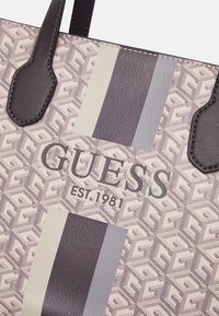 Bolsa tote con un patrón geométrico en beige y marrón, con asas marrón oscuro y el logo embosado "GUESS EST. 1981".