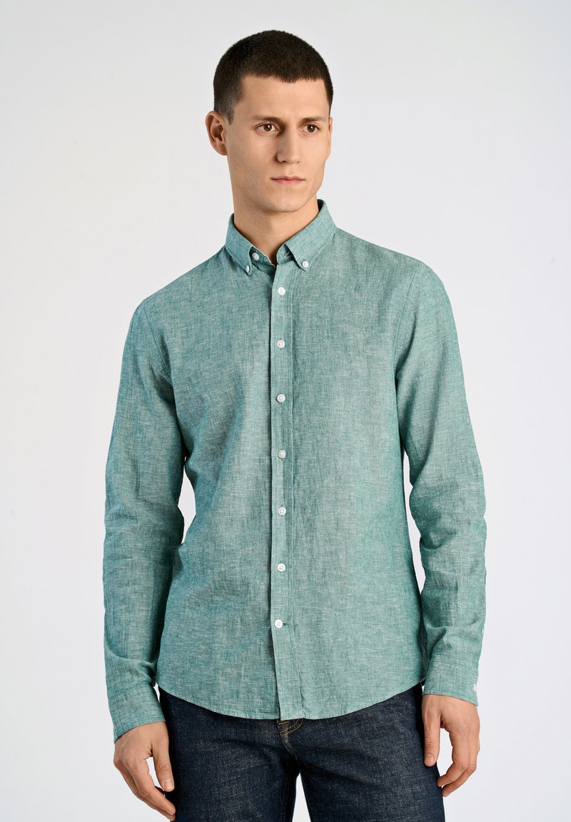 Ljus turkos, knappad skjorta med krage, lång ärm och textilstruktur. Har vita knappar och en rundad nederkant. Bärs med mörka jeans.