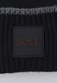 Berretto nero lavorato a maglia con una striscia grigia, dotato di una toppa quadrata nera embosso con la parola "BOSS" in lettere metalliche in rilievo.