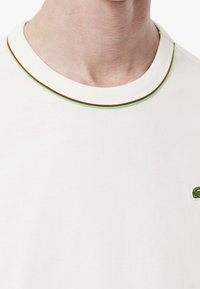 Maglietta bianca a maniche corte con colletto a costine verde e marrone. Presenta un piccolo logo ricamato verde sul petto. Materiale in cotone, texture liscia.