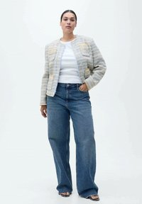 Giacca cropped grigio chiaro con texture, dettagli blu e gialli, abbinata a jeans blu a vita alta e sandali con punta aperta.