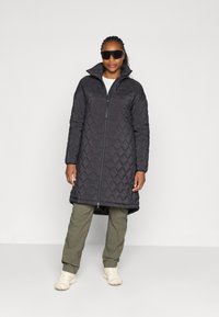 Black quilted long coat with high collar, front zipper, and side pockets, paired with olive green pants and light tan shoes.