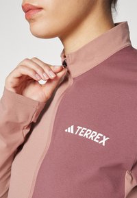 Terrex zip-up jakke i dempede rosa nyanser med en jevn tekstur. Har en svart glidelås og hvit merkevare på brystet.