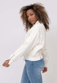 Veste bomber blanche avec fermeture éclair à l'avant, poignets côtelés et zips latéraux, fabriquée dans un tissu lisse avec un léger éclat.