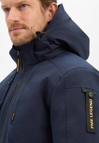 Marineblauwe jas met een hoge kraag, verstelbare capuchon en een zak met een zwart stoffen label met de tekst "PME LEGEND" in het geel.