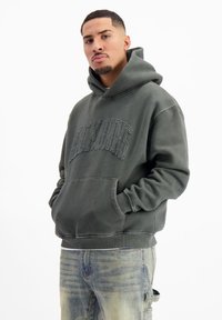 Grijze pullover hoodie met een voorzak, ribgebreide manchetten en zoom. Geborduurde tekst op de borst voegt textuur toe. Draag met een lichte jeans.