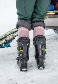 Scarponi da sci neri con accenti verdi e diverse fibbie, indossati con calze rosa, in piedi sulla neve con gli sci visibili sullo sfondo.