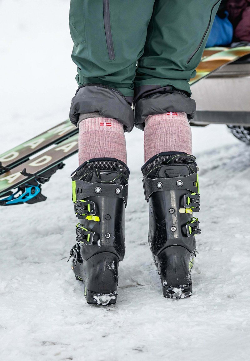 Scarponi da sci neri con accenti verdi e diverse fibbie, indossati con calze rosa, in piedi sulla neve con gli sci visibili sullo sfondo.