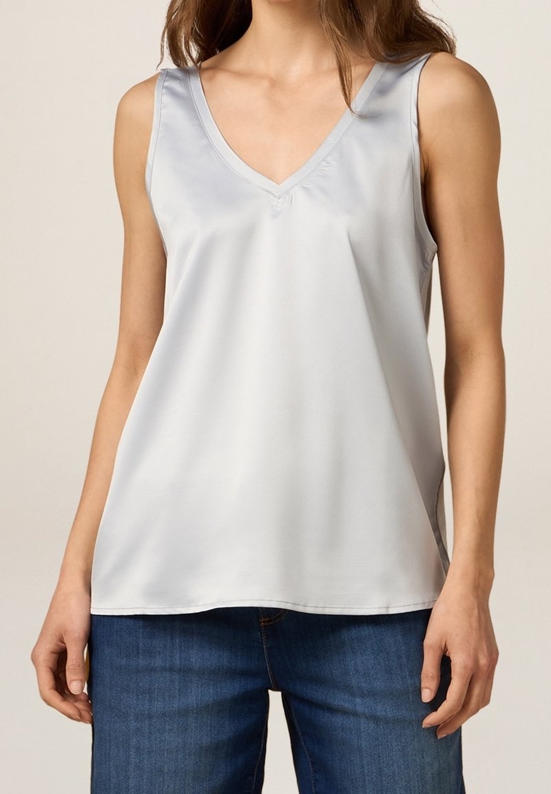 Kvinna klädd i en ärmlös, ljusgrå satin V-ringad topp, tillsammans med blå denimjeans, visad från axlarna till höfterna.