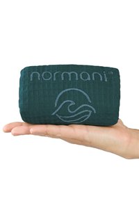 Rechteckiges türkisfarbenes Kissen mit einer strukturierten, gesteppten Oberfläche. Auf der Vorderseite befinden sich ein hellgraues Logo und eine Wellenmarke, das in einer Hand gehalten wird.