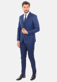 Abito blu navy su misura con vestibilità slim, revers a notched e chiusura a due bottoni. Abbinato a una camicia bianca e una cravatta scura. Scarpe nere lucide.