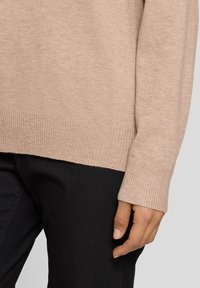 Hellbeiger gestrickter Pullover mit gerippten Bündchen und Saum, kombiniert mit schwarzer Hose. Der Stoff wirkt weich und strukturiert, geeignet zum Schichten.