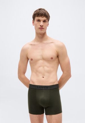 Giovane in piedi contro uno sfondo semplice, indossa boxer Levi's verde scuro con una fascia in vita nera.