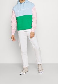 Felpa con blocchi di colore che presenta sezioni blu, rosa e verde, con una tasca frontale e un logo cursivo bianco. Abbinata a pantaloni bianchi e sneakers beige.