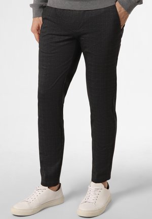 Mężczyzna w czarnych, dopasowanych spodniach o fakturze, z rękami w kieszeniach i białych, niskich trampkach na jasnym tle.