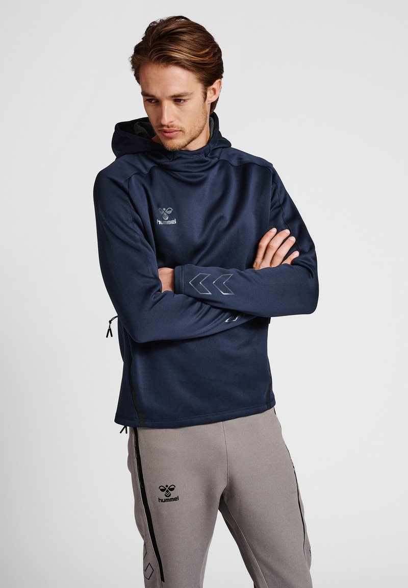 Granatowy hoodie z gładkiego materiału z przednią kieszenią, czarnymi akcentami i srebrnym logo. Posiada kaptur oraz długie rękawy z detalami mankietów.